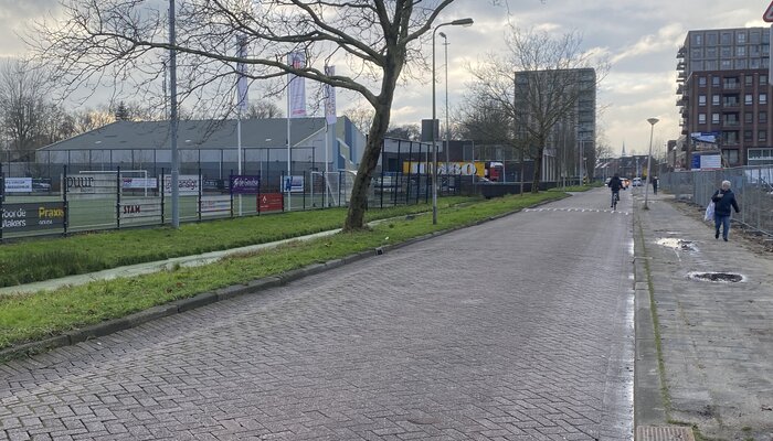 Een straat met klinkers met aan de linkerkant sportvelden. Rechts loopt een voetganger op het betegelde voetpad